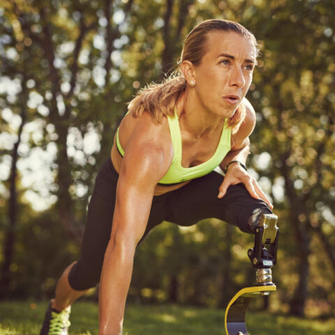 Sportlerin mit einer Beinprothese in der Startposition für einen Sprint auf einer grünen Wiese.