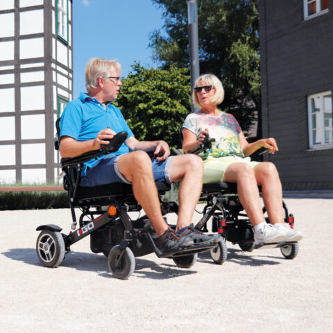 Zwei Personen sitzen in Elektrorollstühlen und unterhalten sich im Freien.