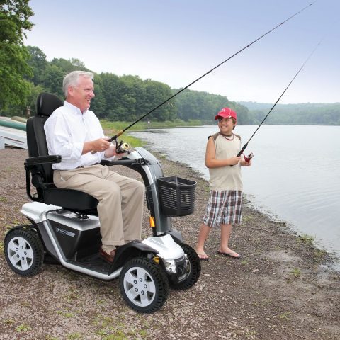 Ein älterer Mann sitzt in einem Elektrorollstuhl am Wasser, während ein Junge neben ihm angelt.