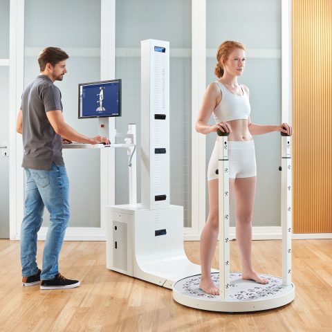 Körperanalysegerät Bauerfeind® BODYTRONIC® 610 mit einer stehenden Person und einem Bediener, der am Computer arbeitet, zur Durchführung von Messungen.